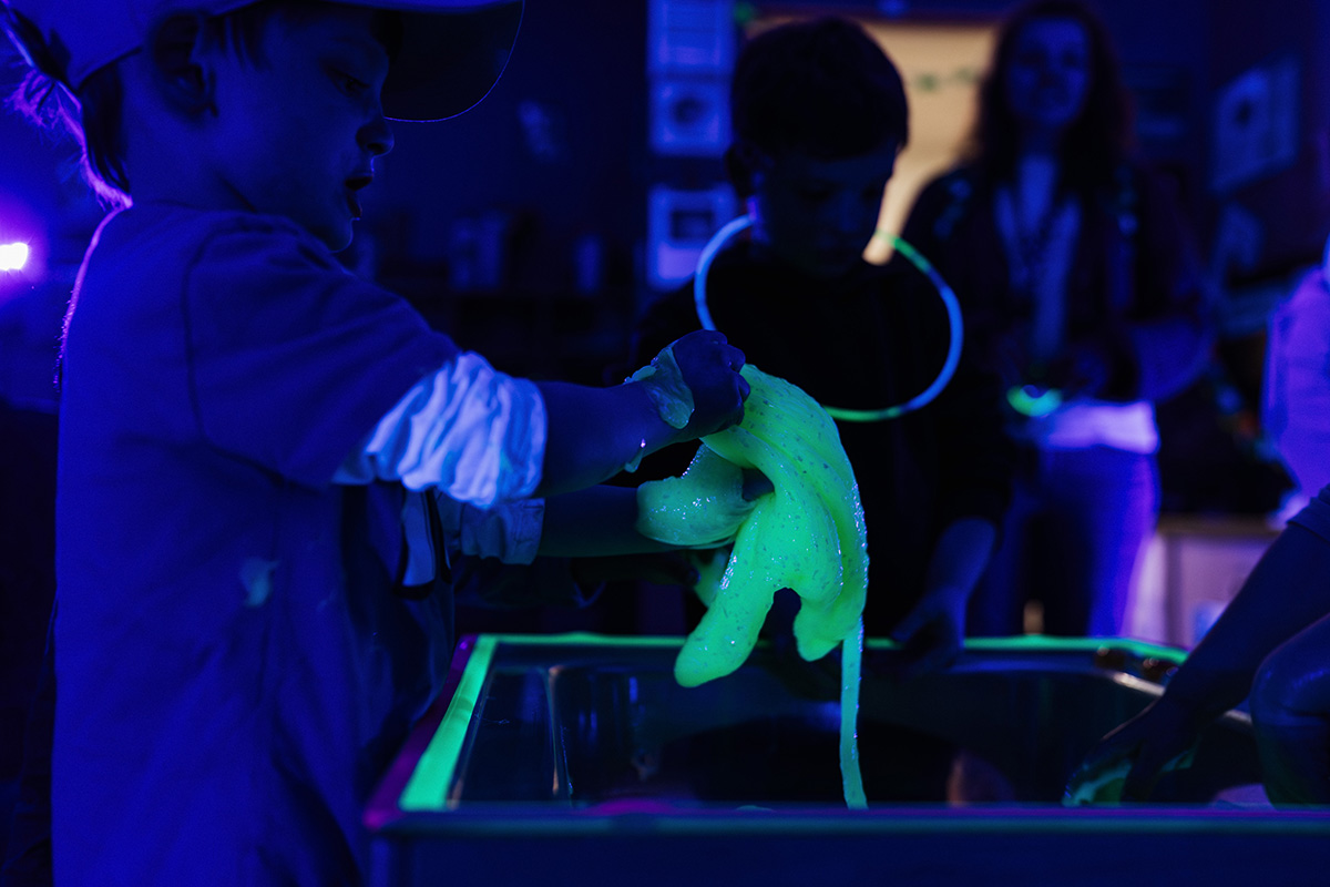 FLINT_Light Up the Night 2026_0006 A child is holding glowing green slime over a container, with a dark background illuminated by blue and purple lights.