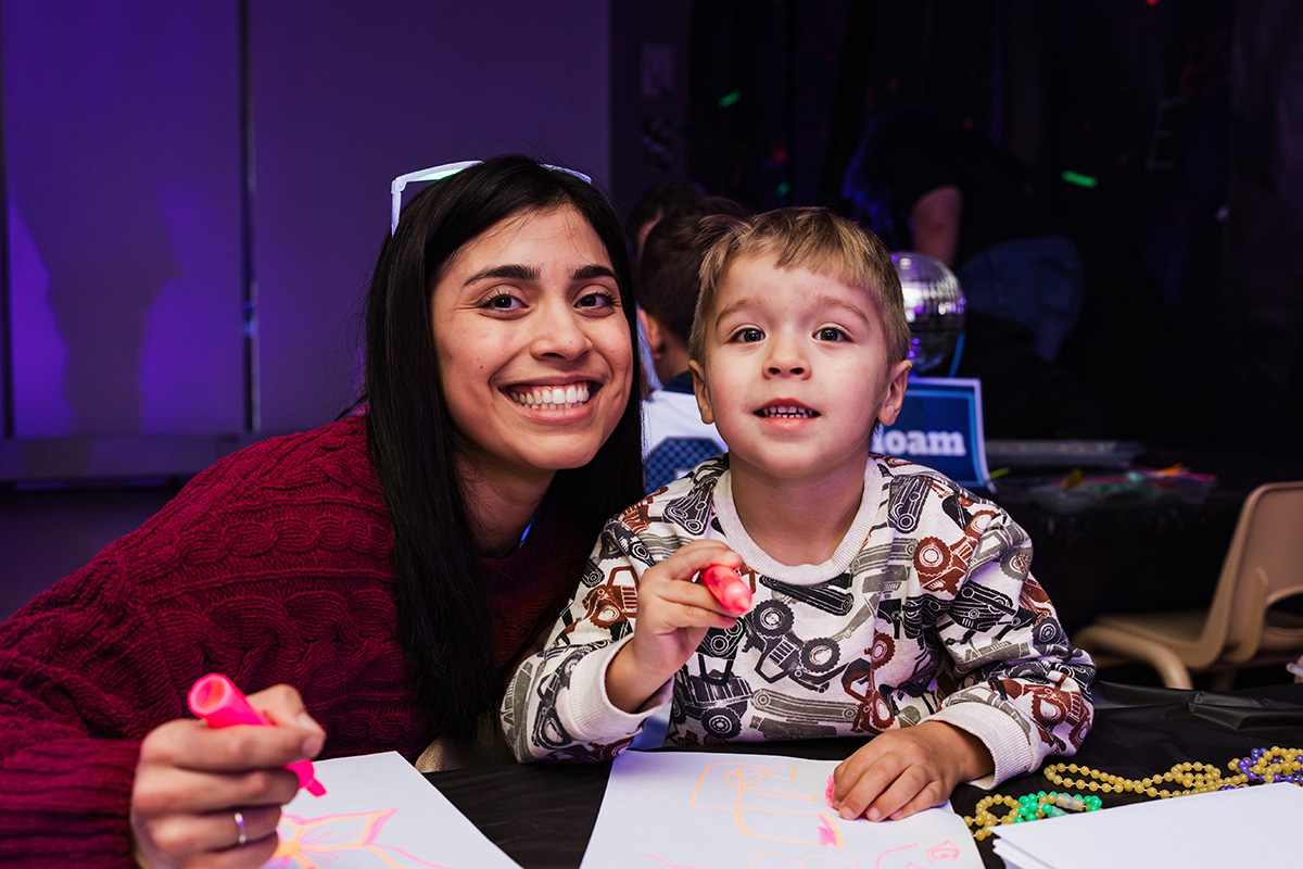 FLINT_Light Up the Night 2026_0009 A person and a child are sitting at a table with drawing materials, creating art on paper. The background features colorful lighting.