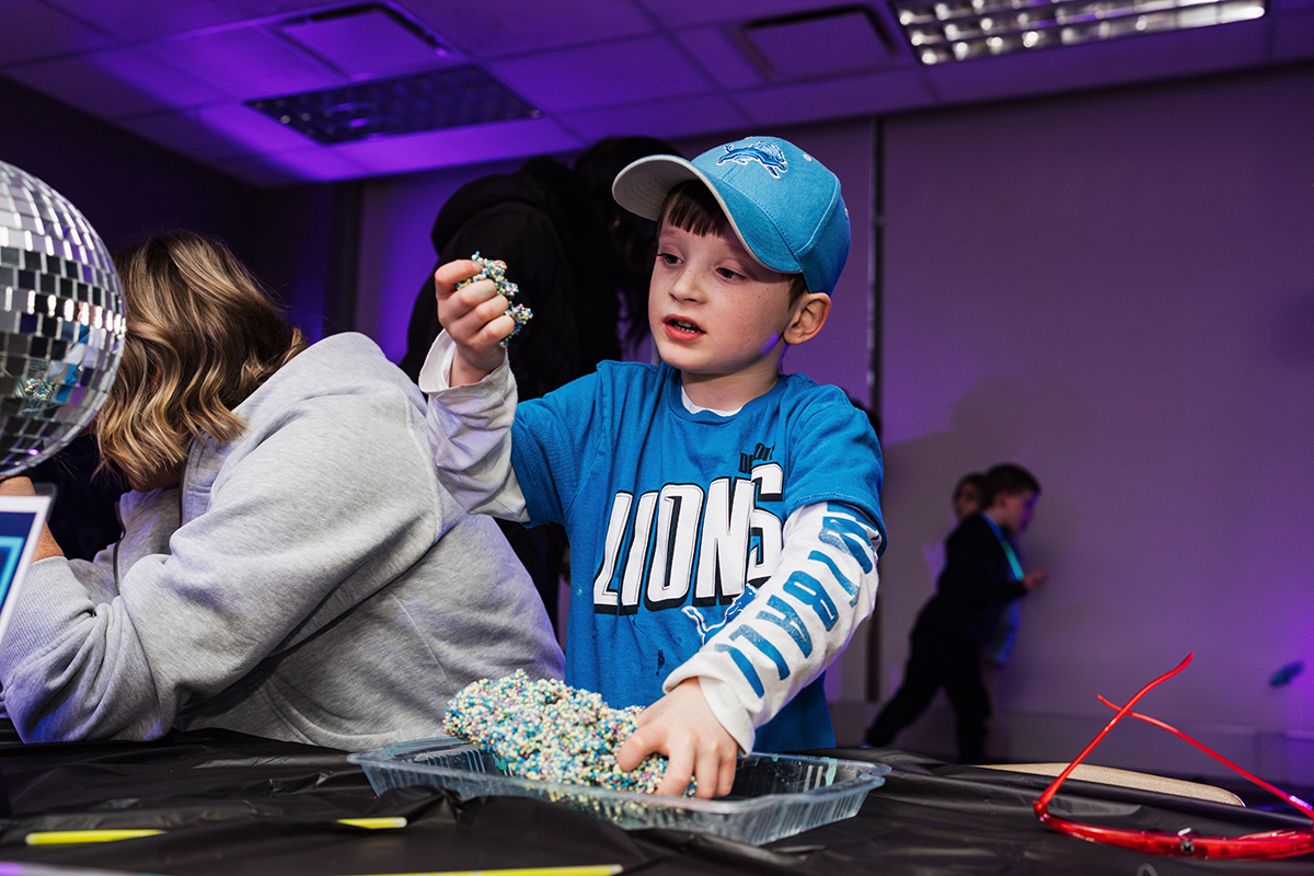 FLINT_Light Up the Night 2026_0012 A child wearing a blue shirt and cap is holding colorful beads while standing at a table with a disco ball in the background.