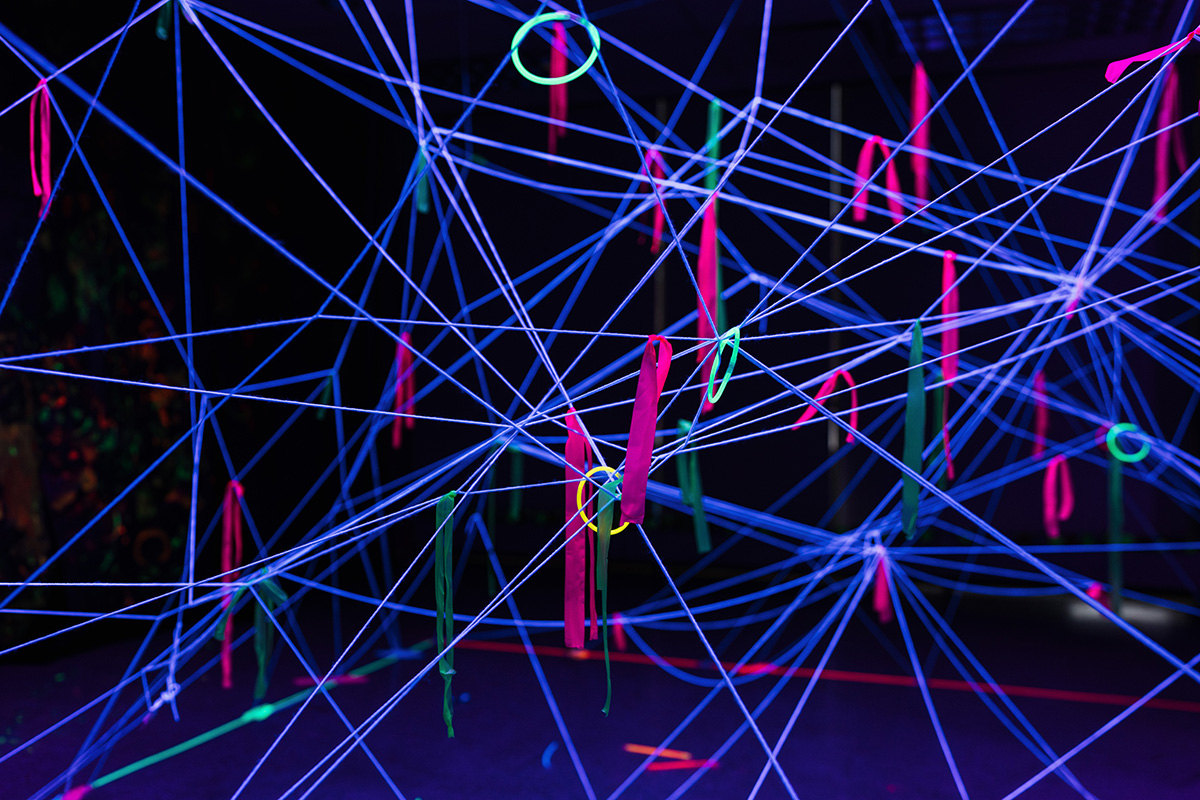 FLINT_Light Up the Night 2026_0014 An abstract installation featuring colorful strings and ribbons in a dark space, with neon pink, green, and blue elements creating a web-like structure.