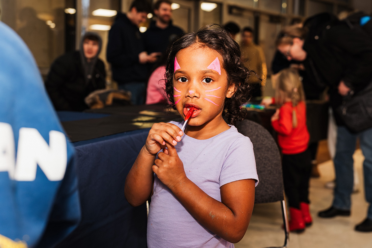 FLINT_Light Up the Night 2026_0019 A young child with curly hair stands in a crowded indoor space, holding their hands together. In the background, several people are engaged in conversation and activities.