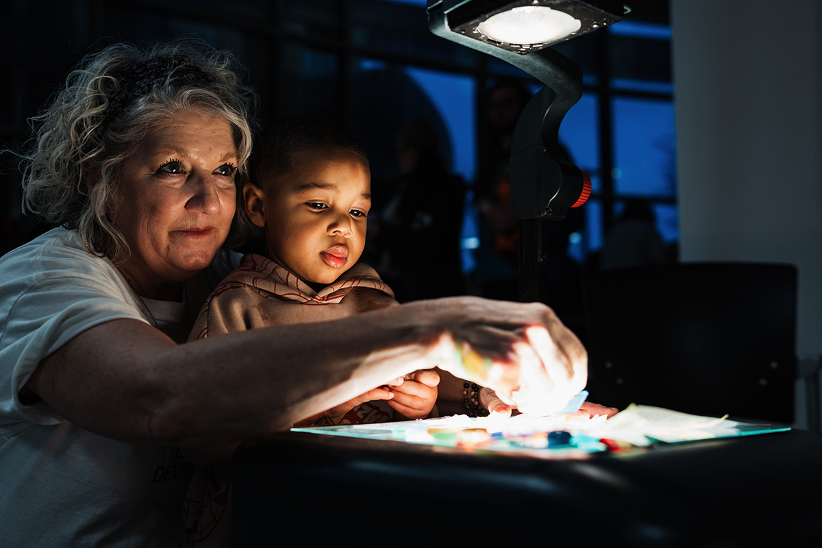 FLINT_Light Up the Night 2026_0024 A person and a child are engaged in an activity at a table illuminated by a lamp, with colorful materials spread out in front of them.