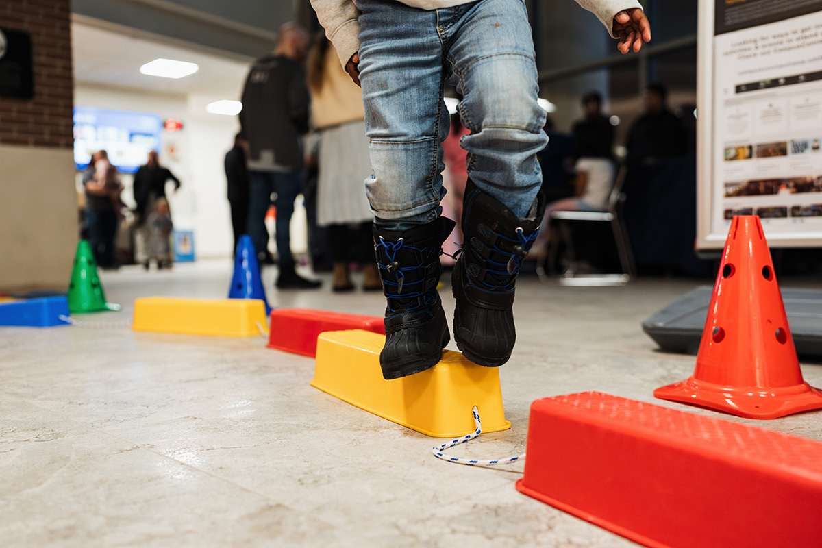 FLINT_Light Up the Night 2026_0026 A child is balancing on a yellow block while stepping over colorful blocks and cones in a busy indoor space.
