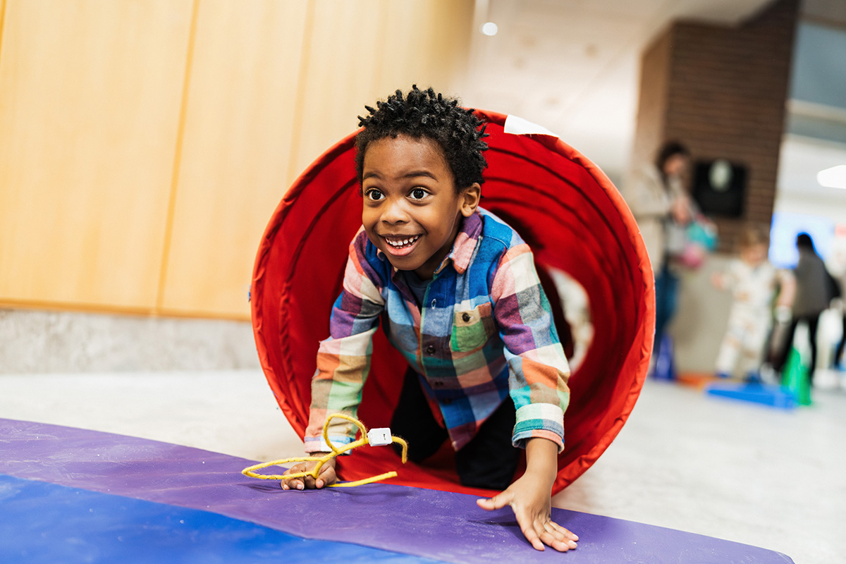 FLINT_Light Up the Night 2026_0028 A child in a colorful plaid shirt is crawling through a red tunnel on a purple mat in a play area.