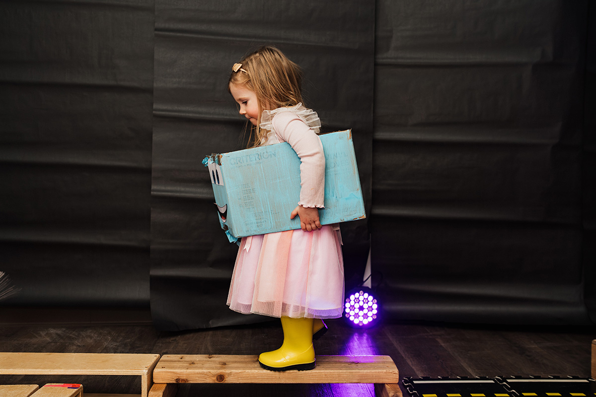 FLINT_Light Up the Night 2026_0035 A child in a pink dress and yellow boots stands on a wooden platform, holding a blue box with a smiling face design.