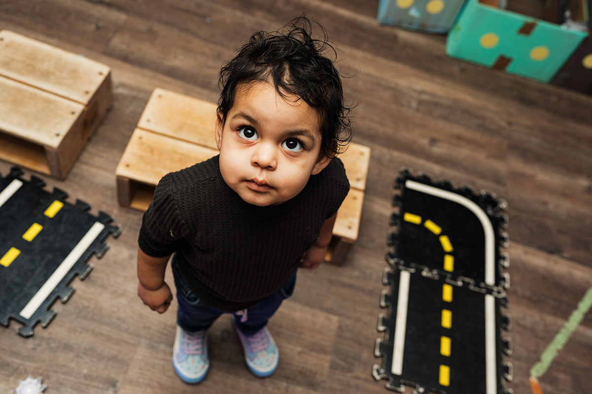 FLINT_Light Up the Night 2026_0036 A child stands on a play mat with road patterns, surrounded by wooden blocks in a playroom setting.