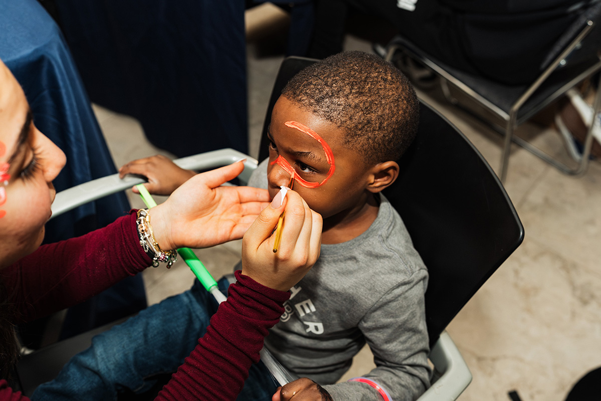 FLINT_Light Up the Night 2026_0054 A child sitting in a chair is having their face painted by an adult, who is holding a paintbrush.