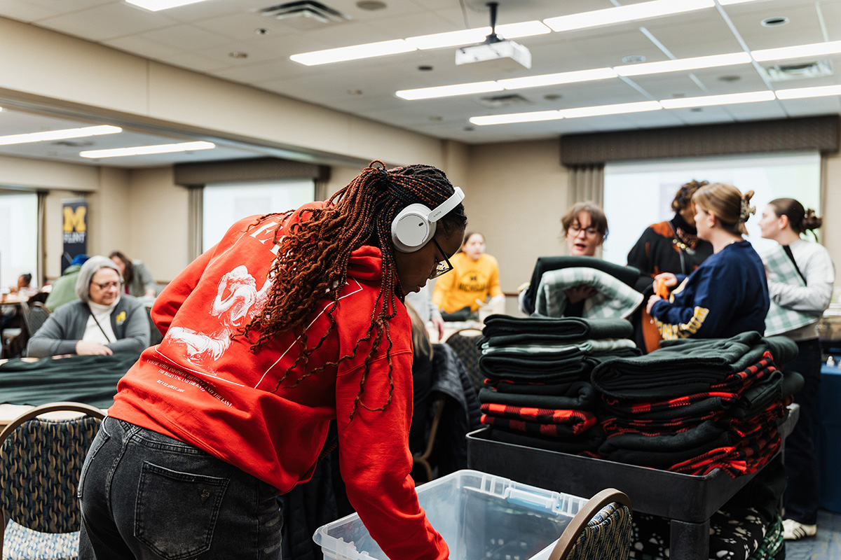 A person in a red sweatshirt is bending over a clear container, while others are gathered around stacks of folded blankets in a room set up for an event.