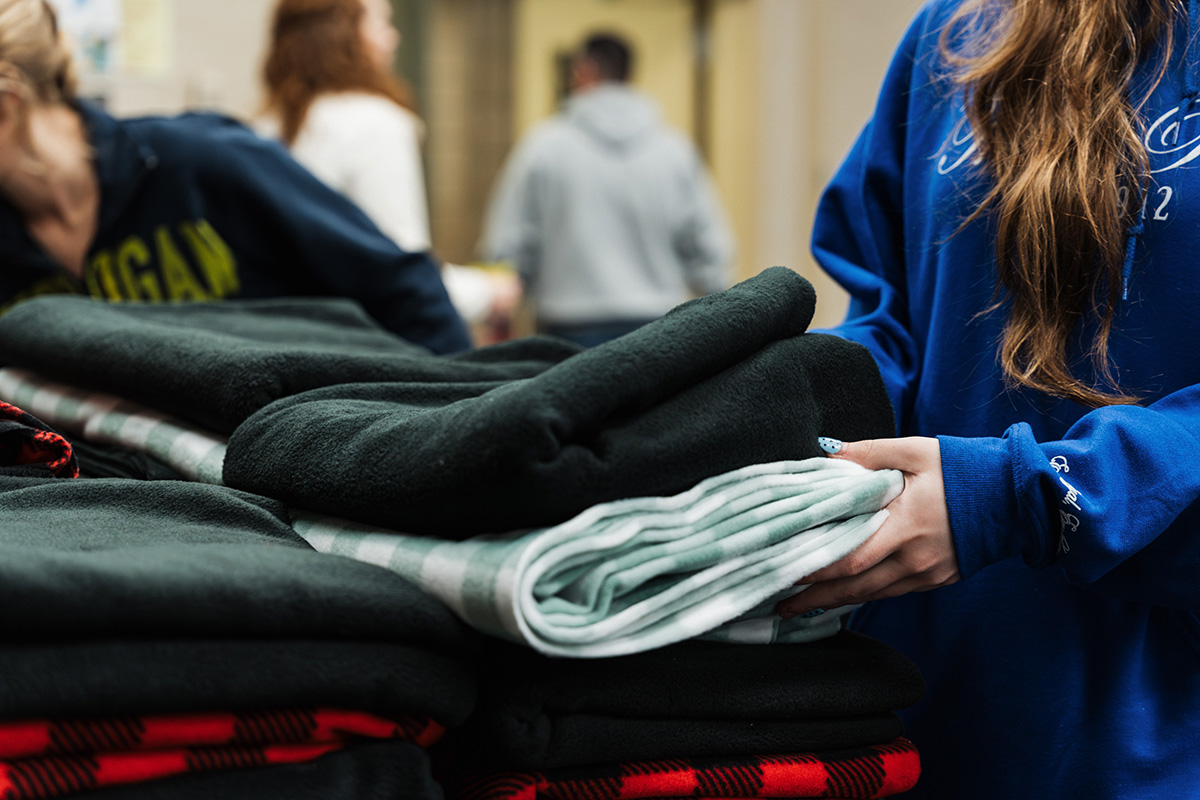 A person wearing a blue hoodie is holding a folded light green blanket while standing next to a stack of various blankets.