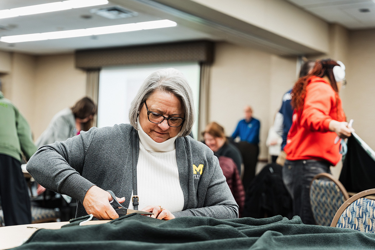A person is cutting fabric with scissors at a table in a room filled with people engaged in various activities.