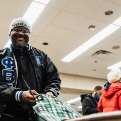 A person wearing a black jacket with blue lettering is holding a pair of scissors and cutting a green and black checkered fabric. In the background, several people are engaged in conversation. The setting appears to be indoors with fluorescent lighting.