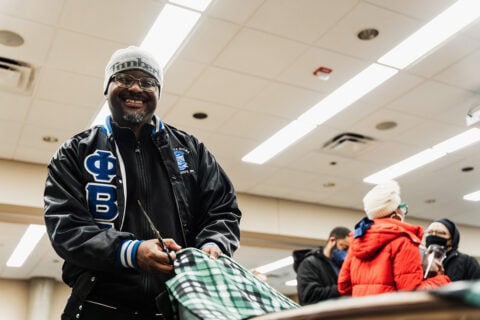 A person wearing a black jacket with blue lettering is holding a pair of scissors and cutting a green and black checkered fabric. In the background, several people are engaged in conversation. The setting appears to be indoors with fluorescent lighting.