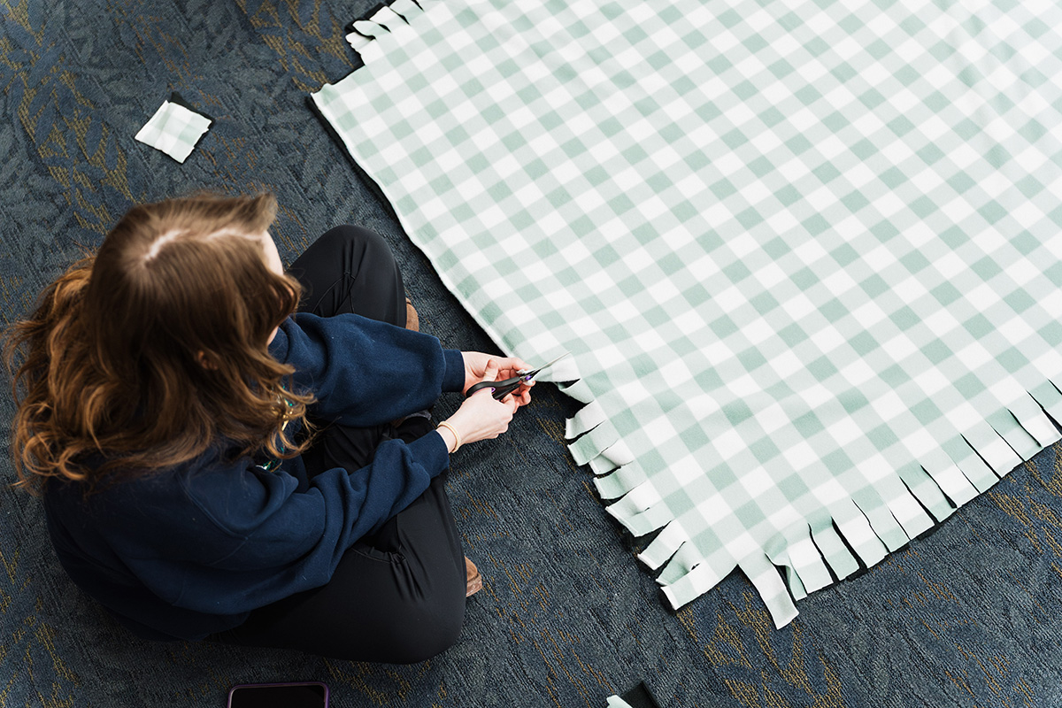 A person sitting on the floor is cutting fringes into a large piece of green and white checkered fabric.