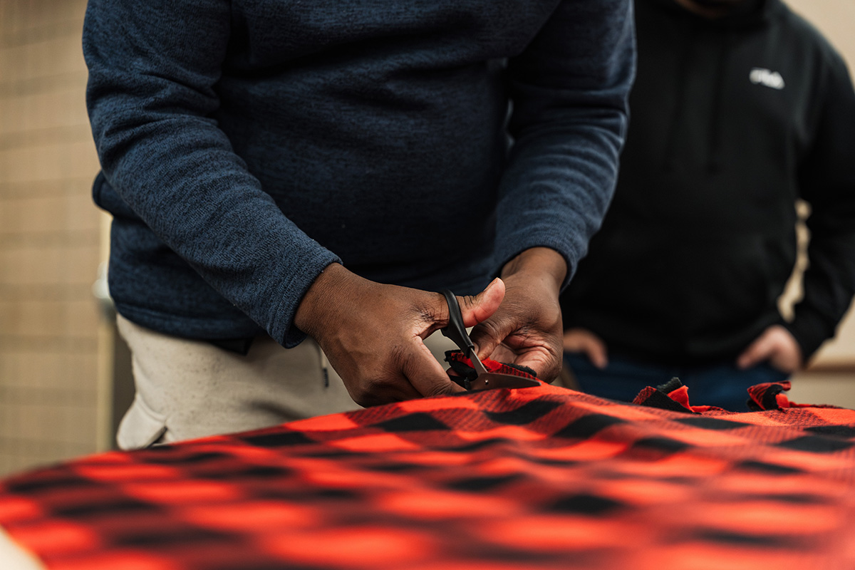 A person is using scissors to cut a piece of red and black checkered fabric, with another person in a black hoodie observing in the background.