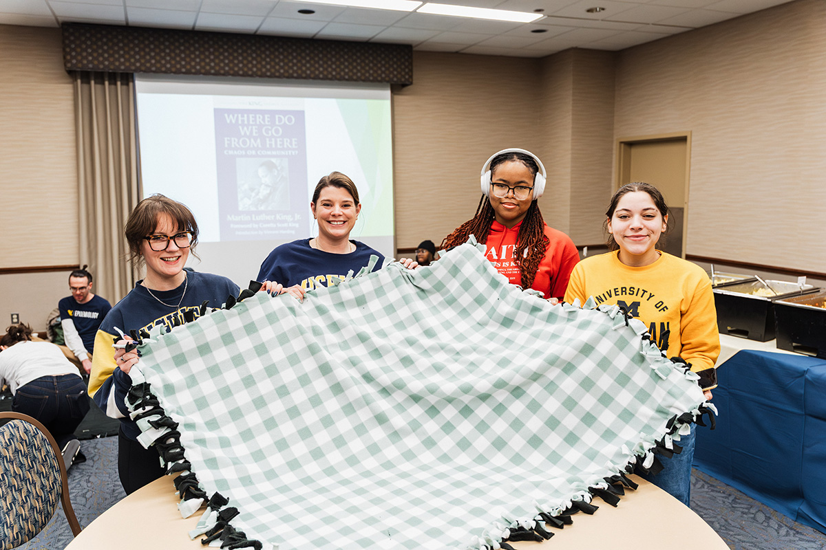 A group of four individuals holds a large green and white checkered blanket in a conference room, with a presentation screen in the background displaying the title "Where Do We Go From Here."