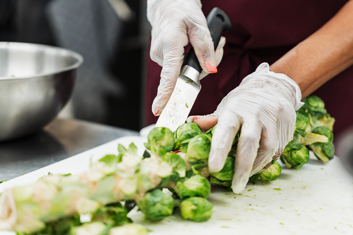A person wearing gloves is chopping Brussels sprouts on a cutting board with a knife, while a metal bowl is visible in the background.