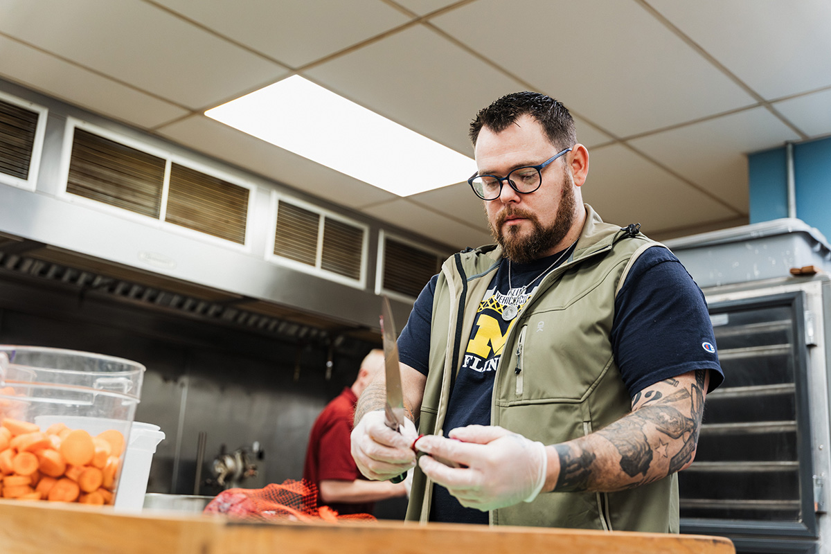 A person wearing a green vest and gloves is holding a knife and preparing food in a kitchen, with a container of chopped carrots nearby.
