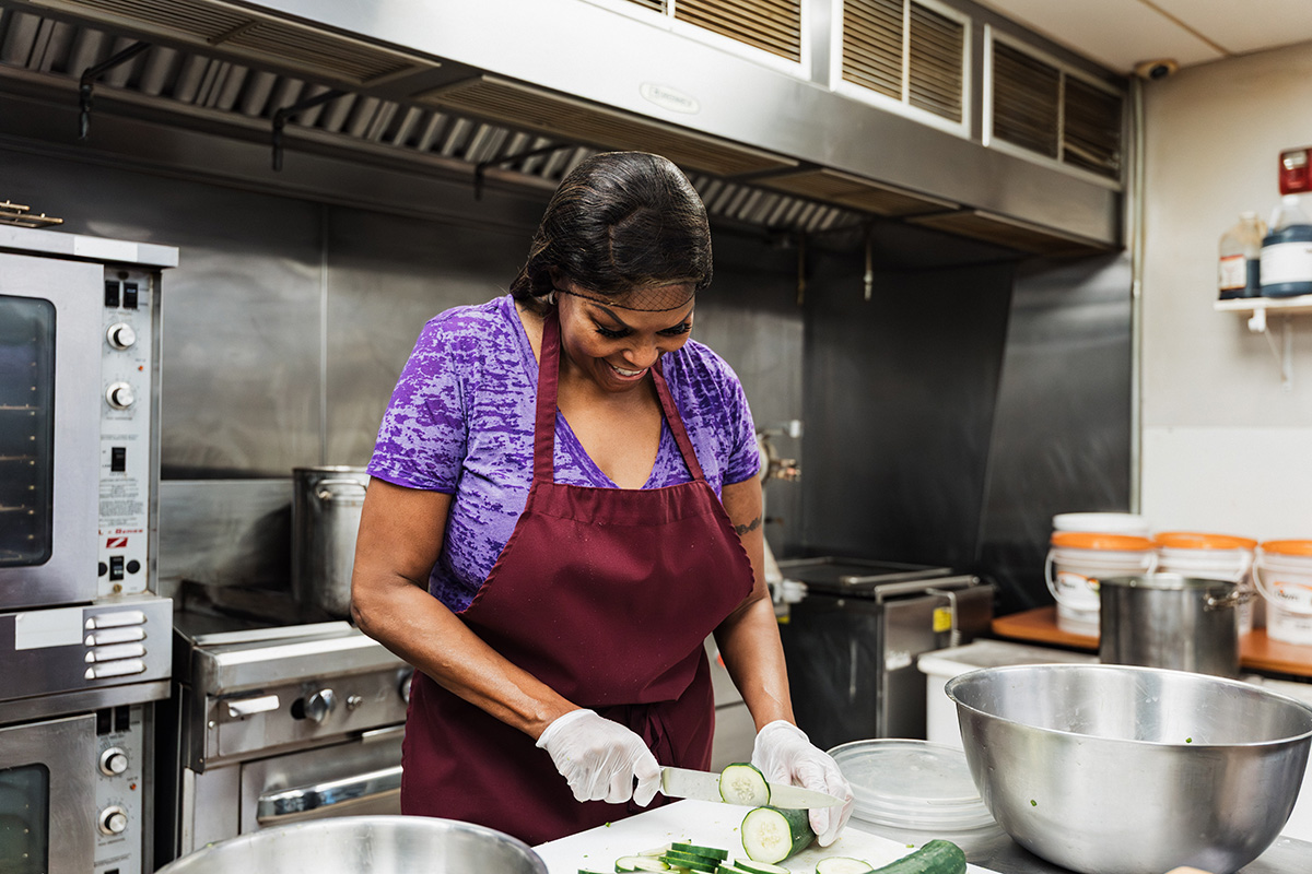 A person in a purple shirt and apron is slicing cucumbers on a cutting board in a kitchen, with various kitchen equipment in the background.