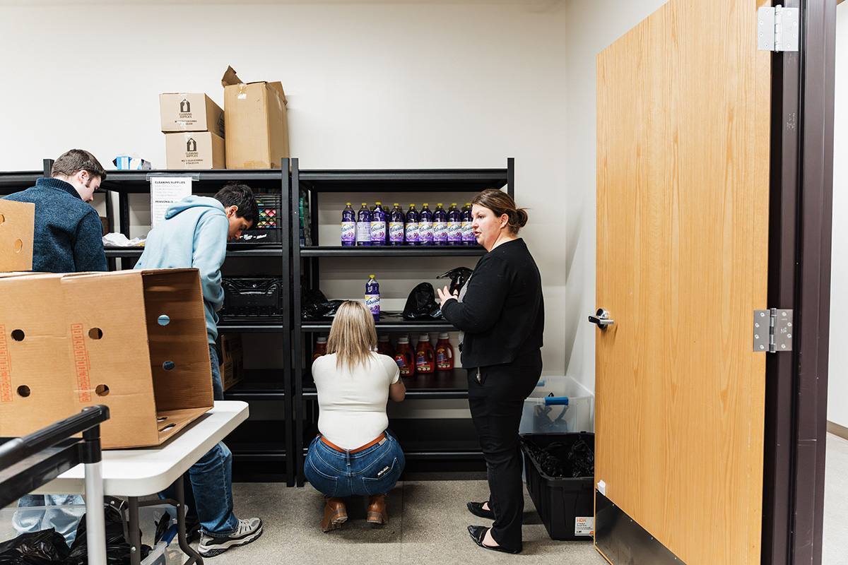 A small storage room with shelves holding various bottles and containers. Two people are organizing items on the shelves, while another person is crouched down, inspecting items. There are cardboard boxes and a door visible in the background.