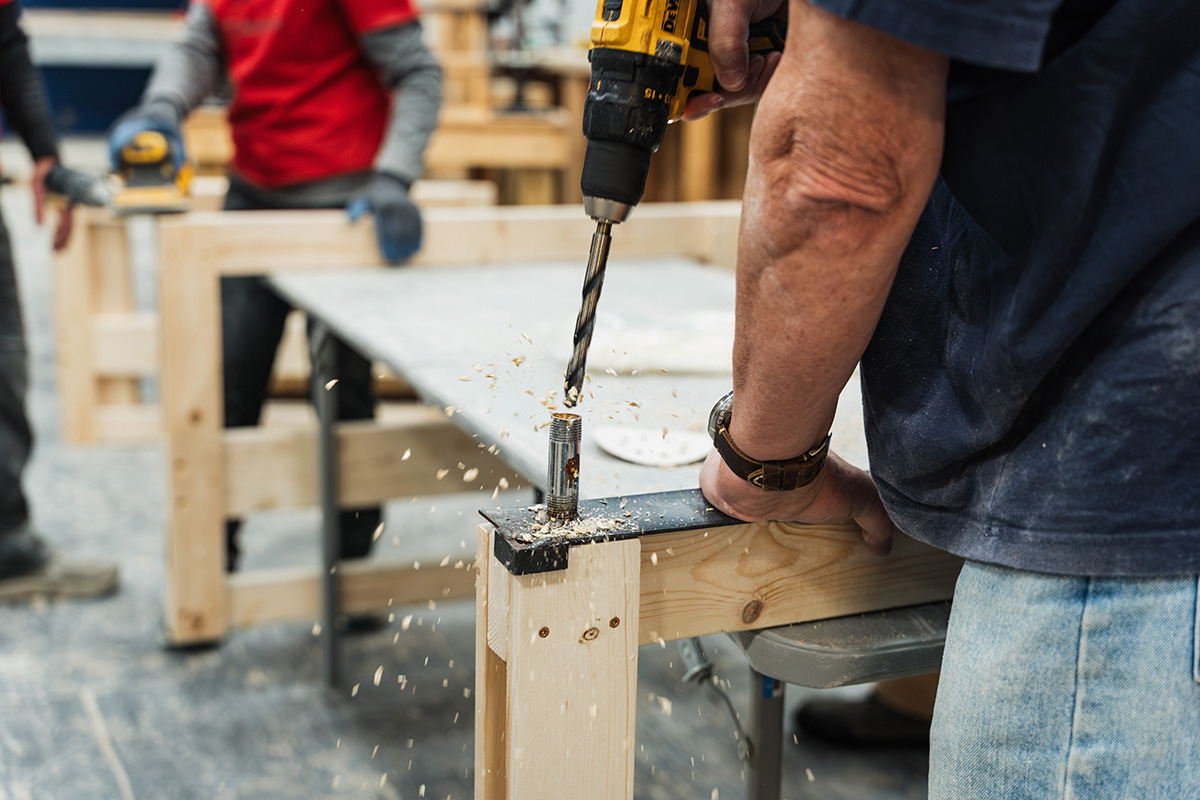 A person is using a power drill to create a hole in a wooden piece, with wood shavings flying from the drill. In the background, two individuals are working on a wooden structure.