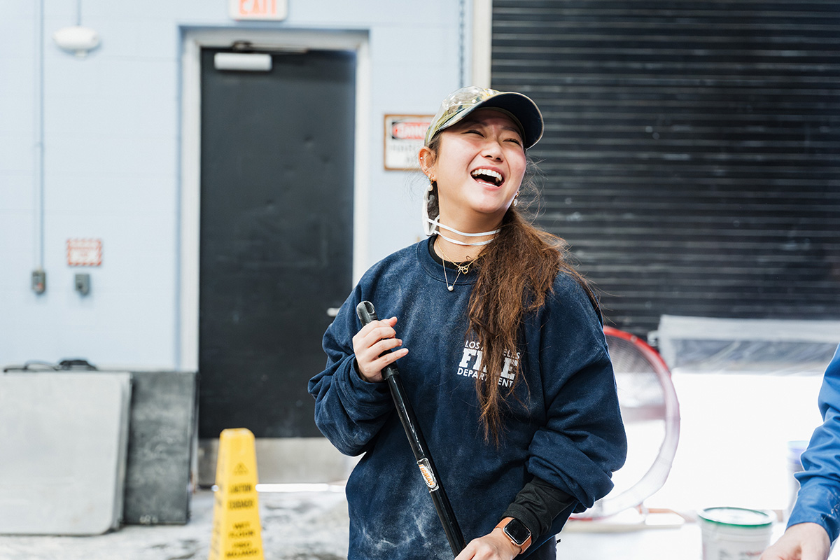 A person wearing a navy sweatshirt with "Los Angeles Fire Department" printed on it, holding a tool, in a room with a black door and caution sign.