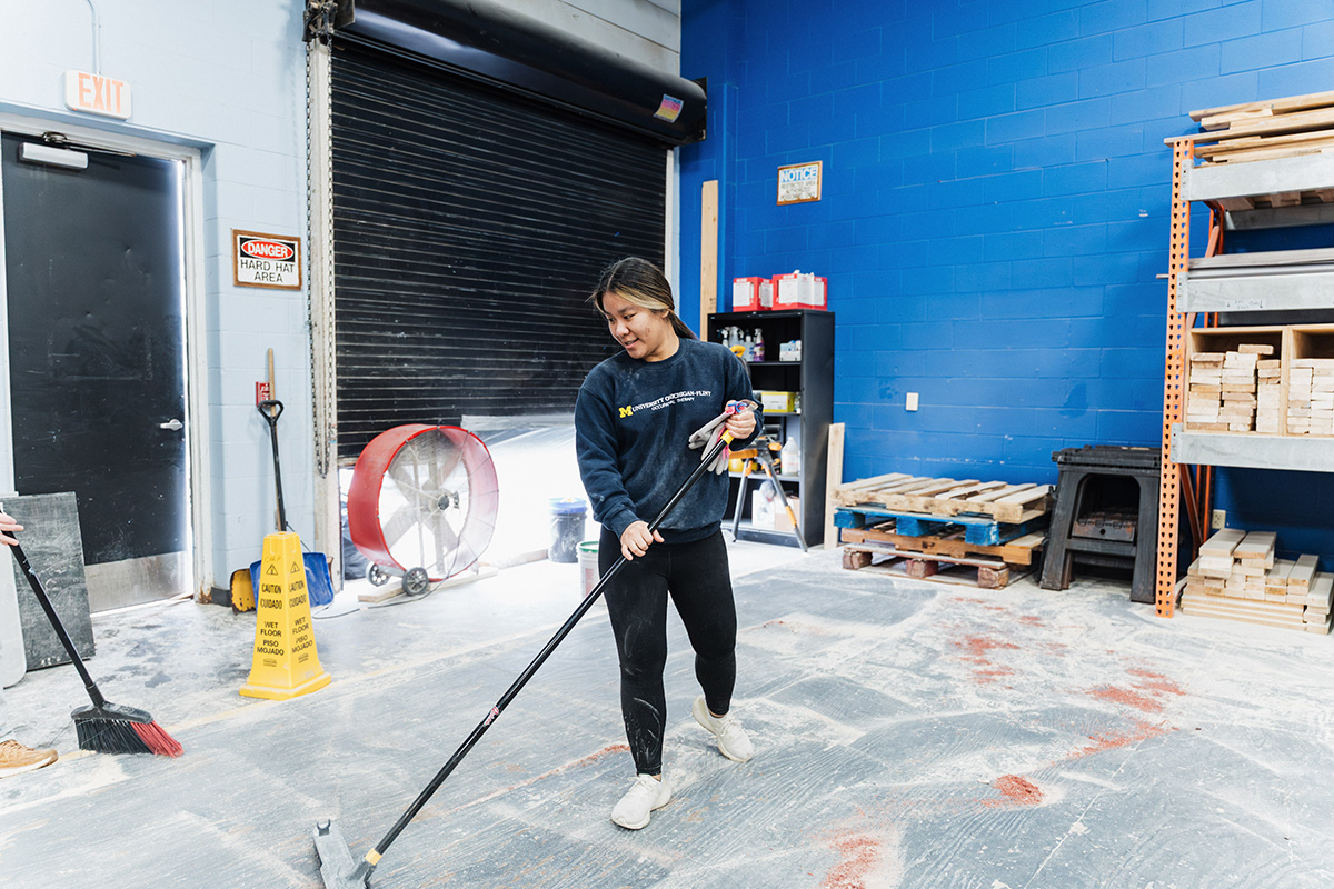 A person is sweeping the floor in a workshop with blue walls. There are shelves with materials and a caution sign nearby.