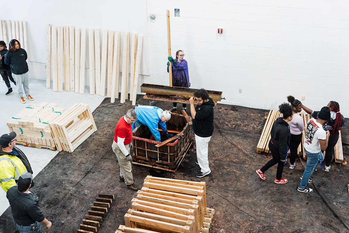 A group of people is engaged in an activity in a large, open space with wooden planks stacked against the walls. Some individuals are working with a large wooden container, while others are observing or interacting nearby.