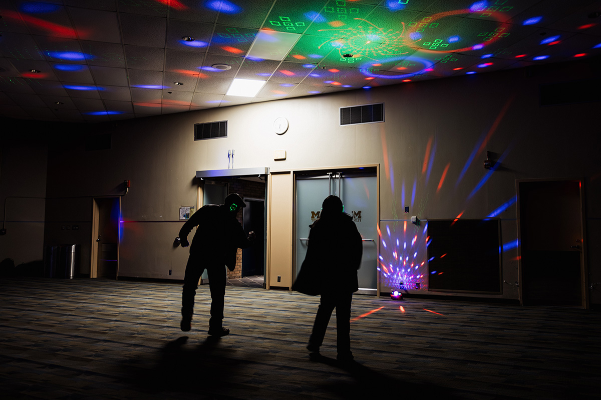 Two silhouetted figures dance in a dimly lit room with colorful lights projected on the ceiling and walls.