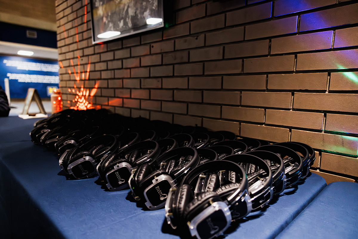 A row of black headphones is neatly arranged on a blue tablecloth against a brick wall, with colorful light reflections in the background.