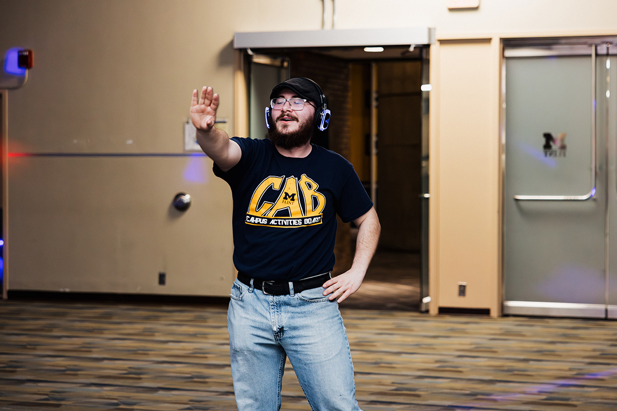 A person wearing a black t-shirt with "CAB" and "CAMPUS ACTIVITIES BOARD" printed on it is standing with one hand raised, wearing headphones, in a spacious indoor area with a patterned floor.