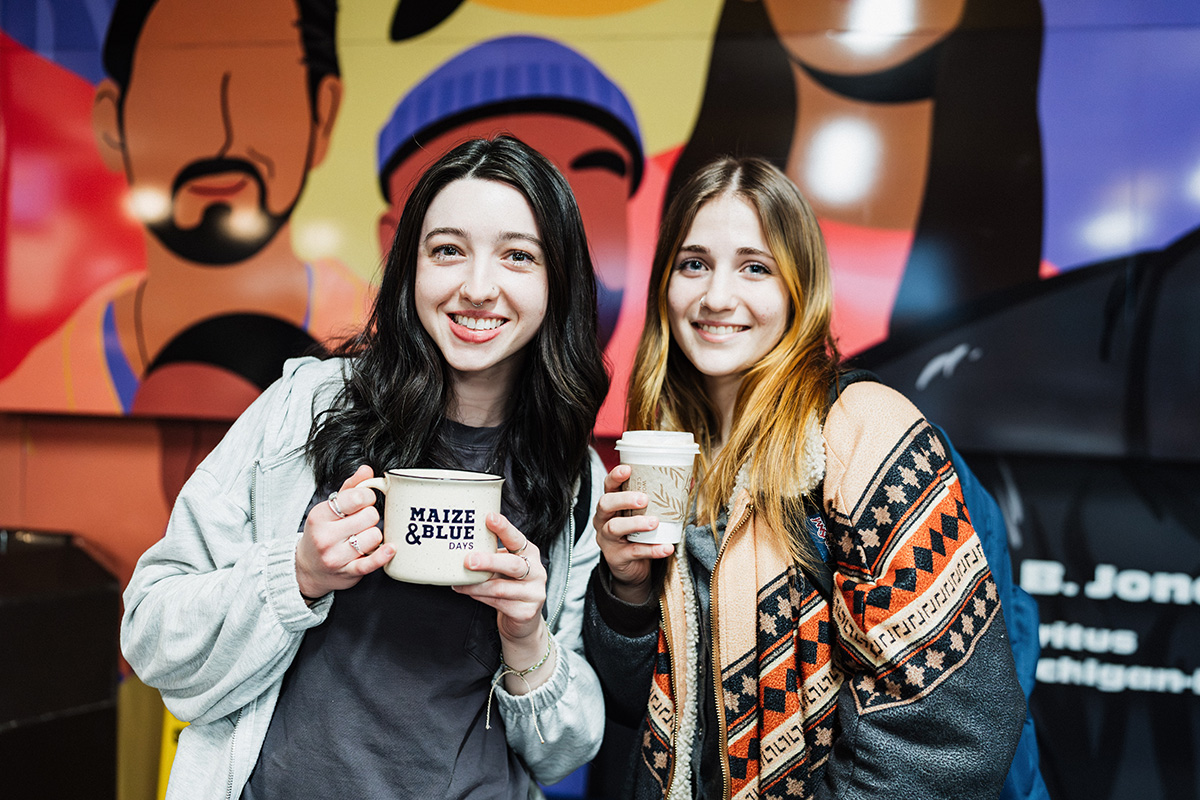 Two individuals are holding coffee cups in front of a colorful mural. One cup has the text "MAIZE & BLUE DAYS." The background features vibrant artwork.