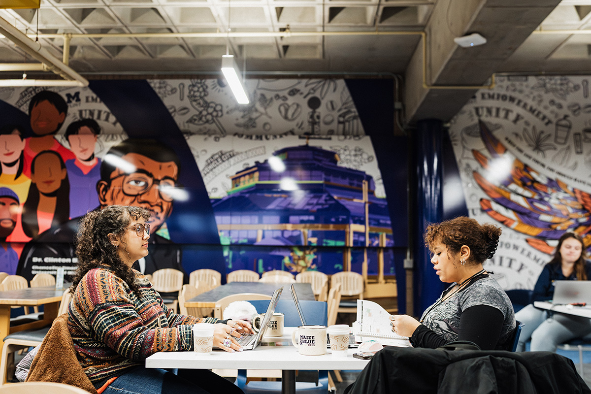 Two people are seated at a table in a café, with laptops and coffee cups in front of them. The background features a colorful mural with various illustrations and text.
