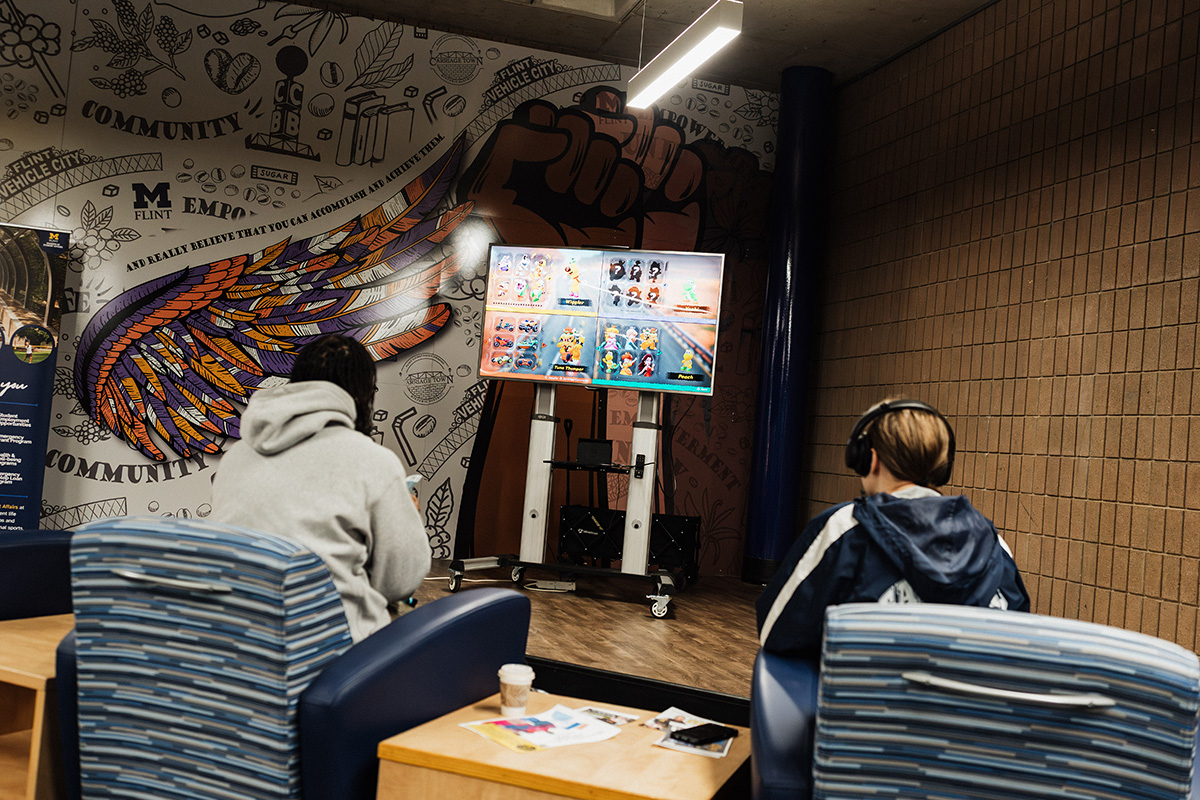Two people are sitting in chairs facing a TV screen displaying a video game. The background features a colorful mural with wings and various text elements. One person is wearing a hoodie, and the other is wearing headphones. A coffee cup and some papers are on a table in front of them.
