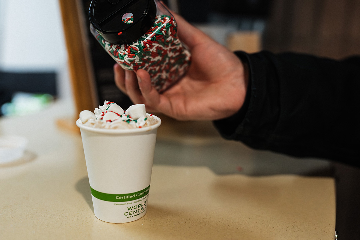 A hand is pouring colorful sprinkles from a container onto a cup filled with whipped cream and marshmallows. The cup is labeled "Certified Compostable" and "WORLD CENTRIC."