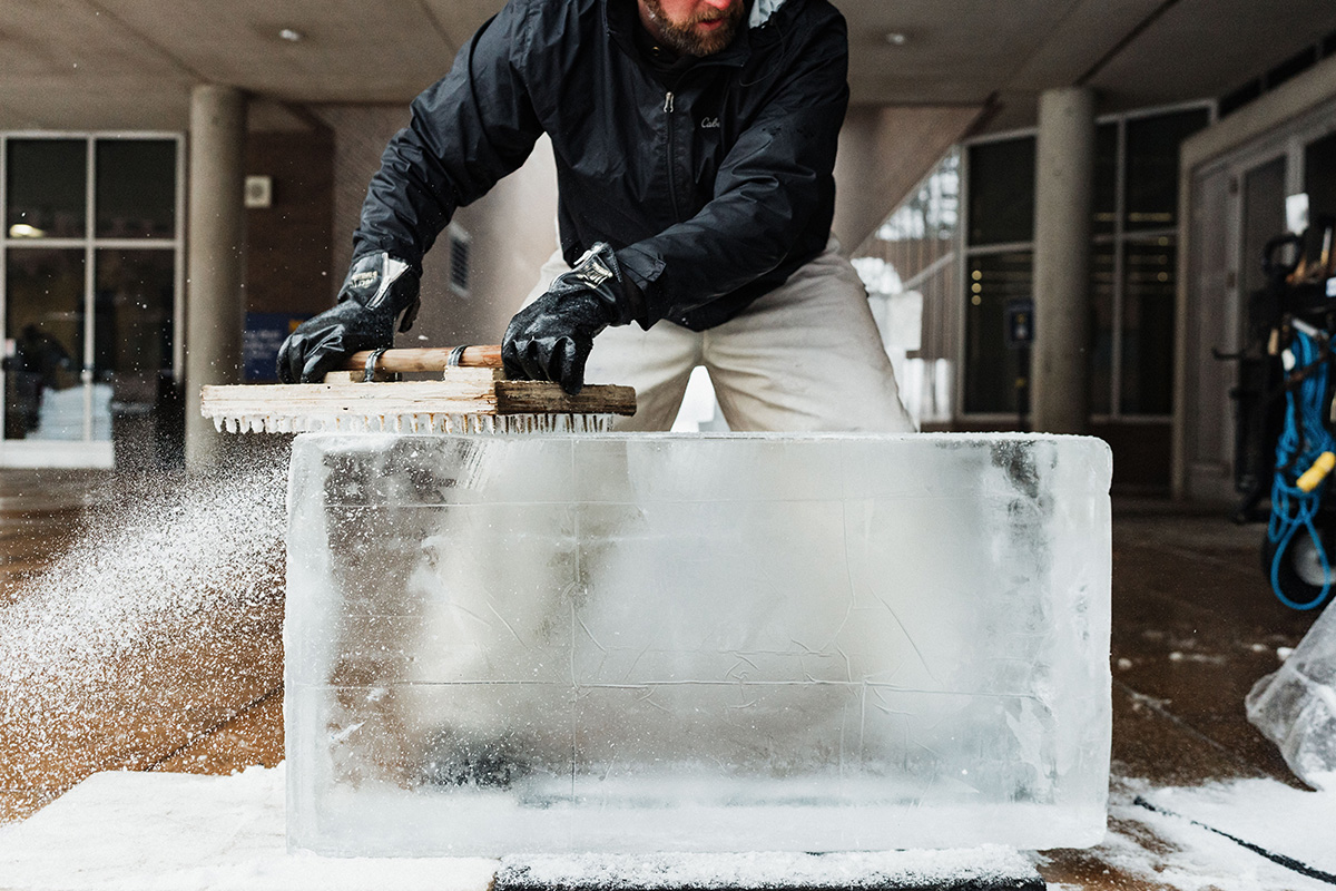 A person in a black jacket and gloves is using a tool to carve a large block of ice, with ice shavings flying off.