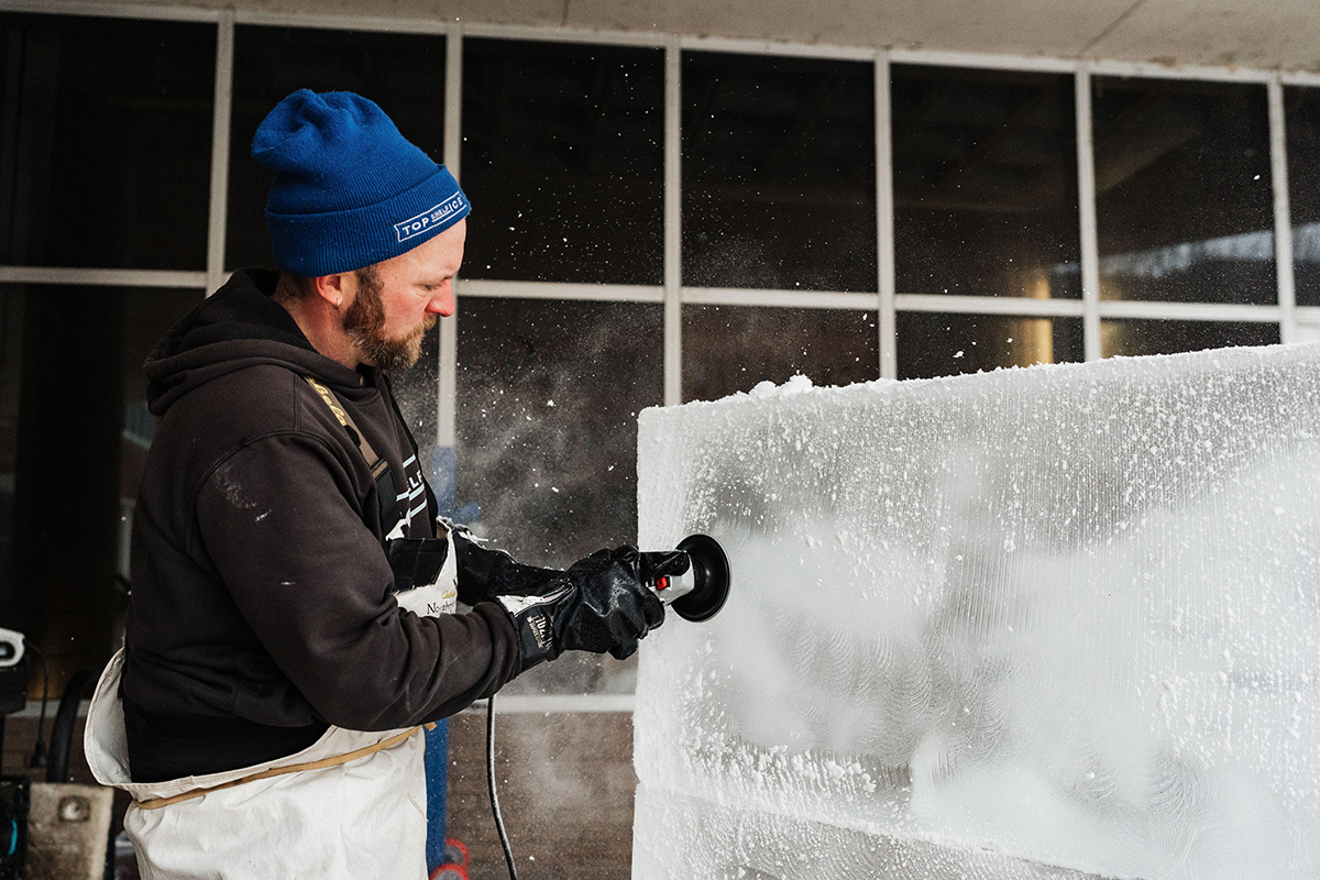 A person wearing a blue beanie and gloves is using a tool to carve an ice block, with ice shavings flying around.