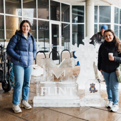 Two people stand in front of an ice sculpture featuring the word "FLINT" and a snowflake design, with a cartoon character nearby. A cart with tools is visible in the background.