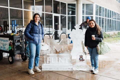 Two people stand in front of an ice sculpture featuring the word "FLINT" and a snowflake design, with a cartoon character nearby. A cart with tools is visible in the background.