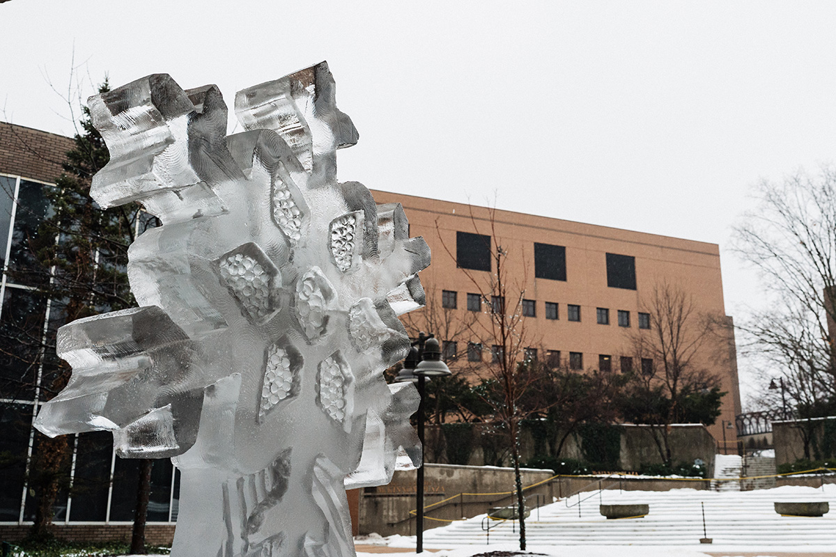 An ice sculpture of a snowflake is prominently displayed in the foreground, with a building and trees in the background. Snow is lightly falling.