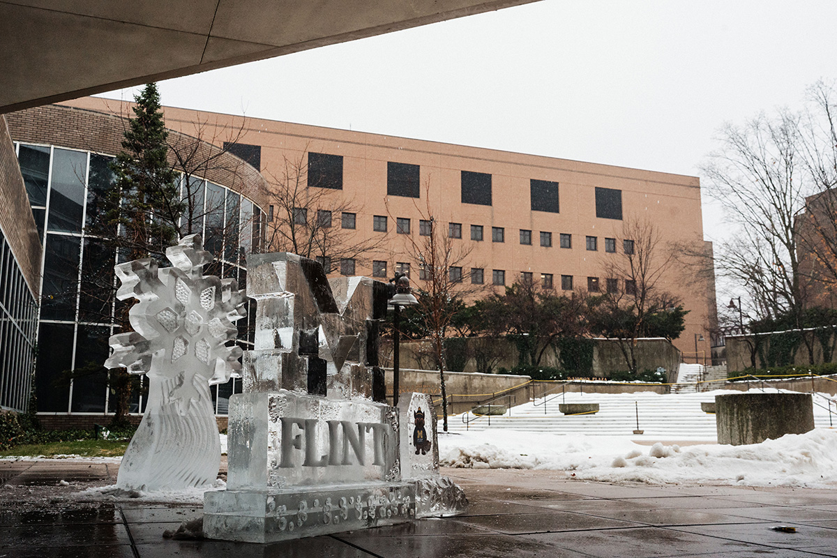 An ice sculpture featuring a snowflake and the word "FLINT" is displayed outdoors, with a building and snow-covered ground in the background.