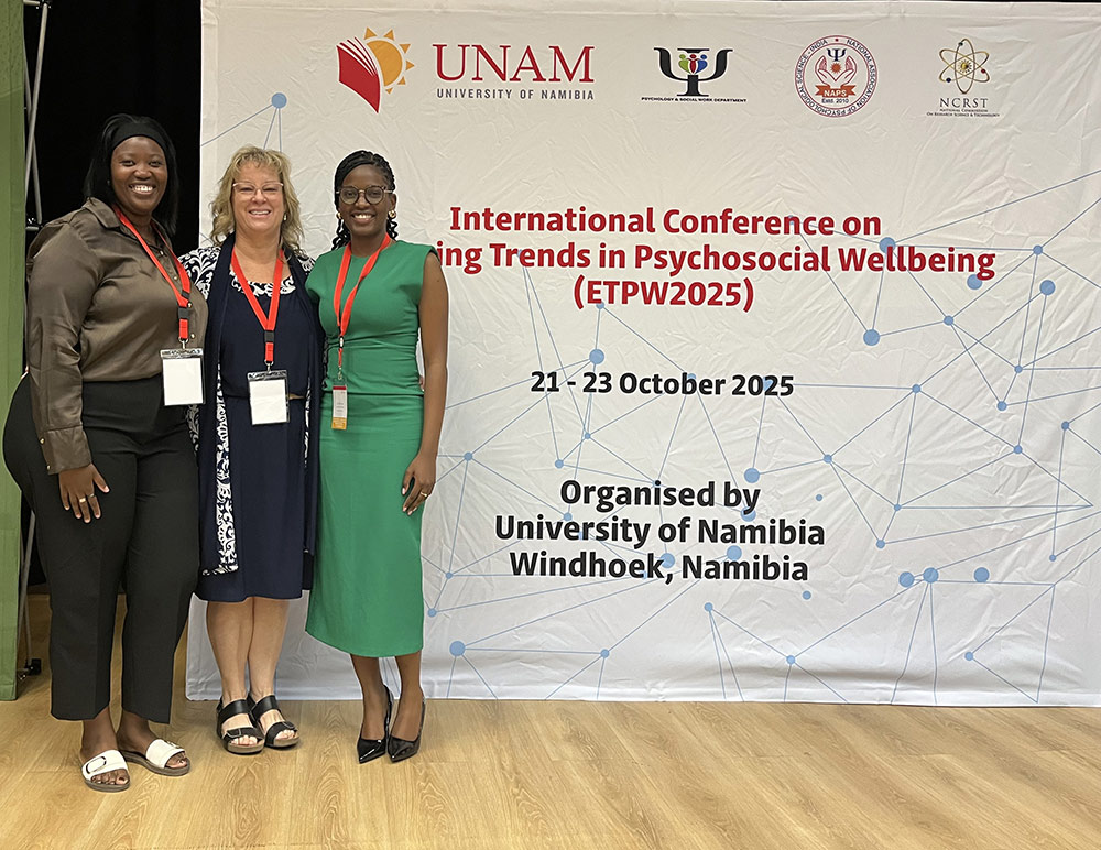 Three women stand in front of a banner for the International Conference on Emerging Trends in Psychosocial Wellbeing (ETPW2025), organized by the University of Namibia, taking place from October 21 to 23, 2025, in Windhoek, Namibia.