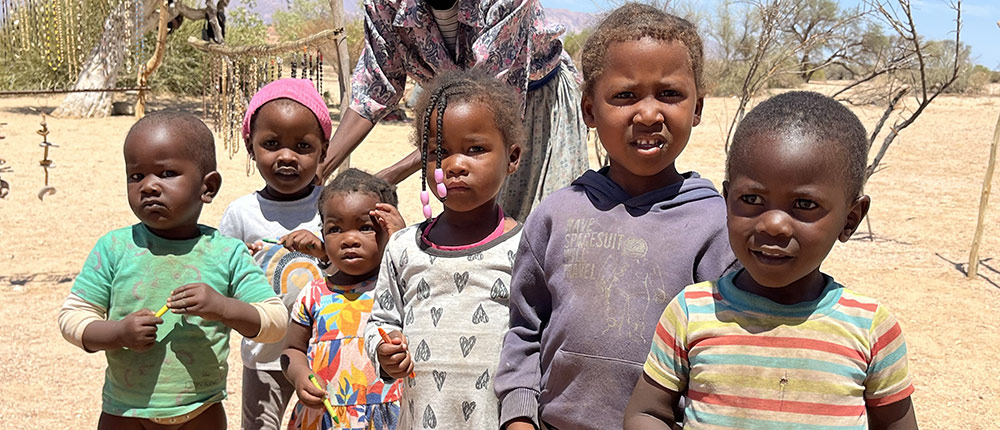 A group of children standing together outdoors, with a woman behind them. The setting appears to be a dry, rural area.