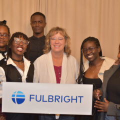 A group of individuals standing together, holding a sign that reads "FULBRIGHT." The background features a textured curtain.