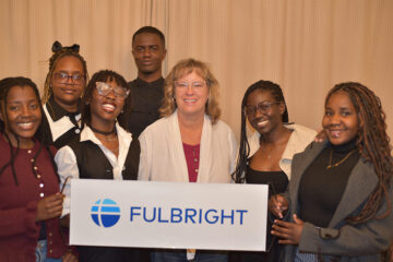 A group of individuals standing together, holding a sign that reads "FULBRIGHT." The background features a textured curtain.