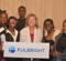 A group of individuals standing together, holding a sign that reads "FULBRIGHT." The background features a textured curtain.