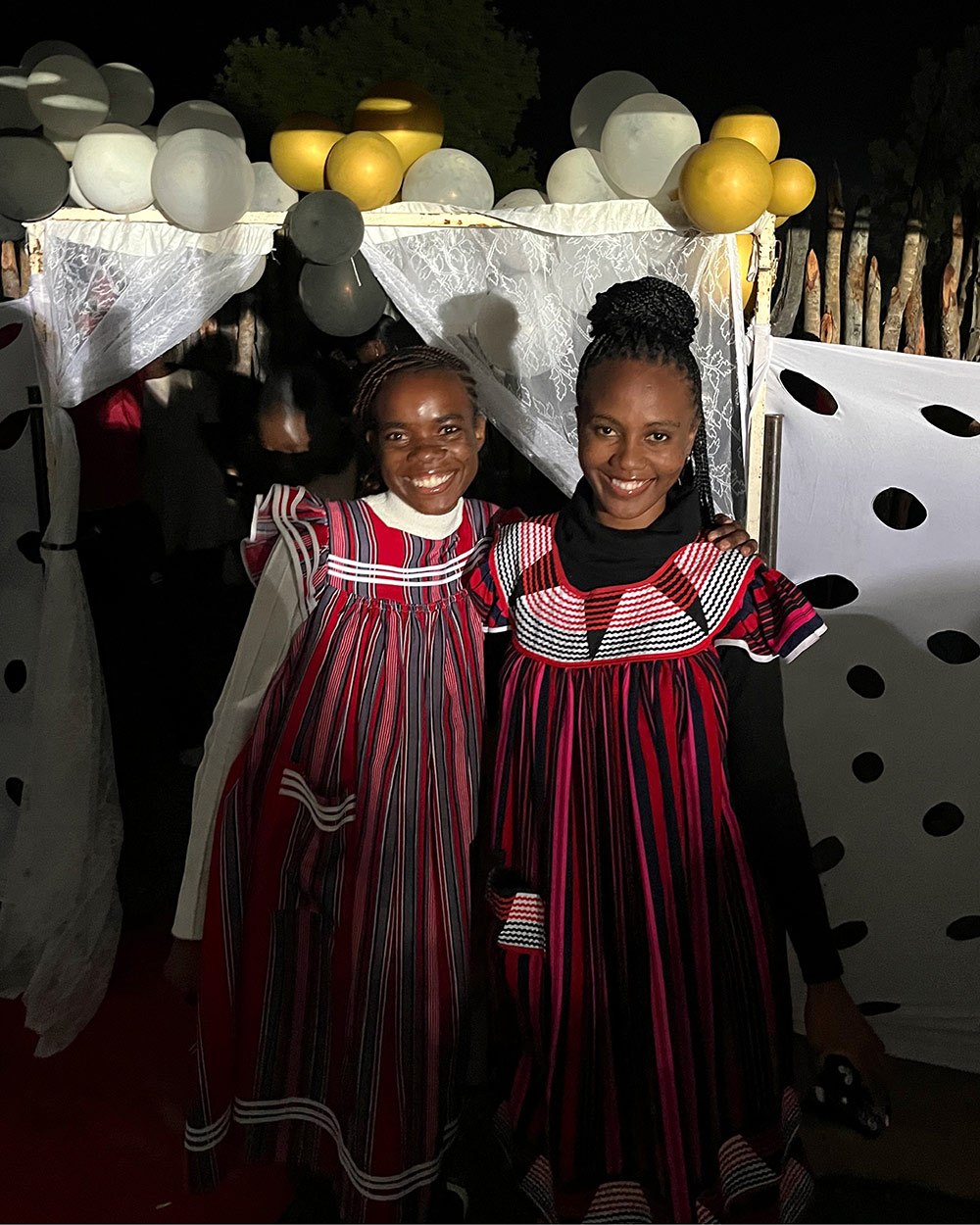 Two individuals wearing traditional red and white dresses stand in front of a decorated entrance with balloons and a festive backdrop.