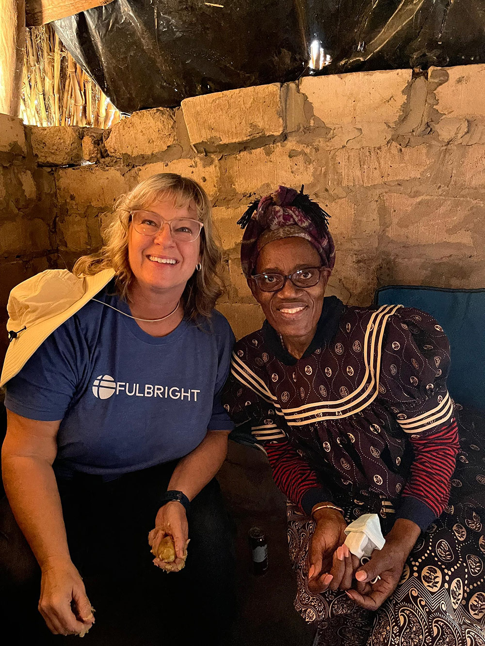 Two individuals are sitting together in a dimly lit room with stone walls. One person is wearing a blue shirt with the word "FULBRIGHT" and the other is dressed in a patterned traditional outfit.