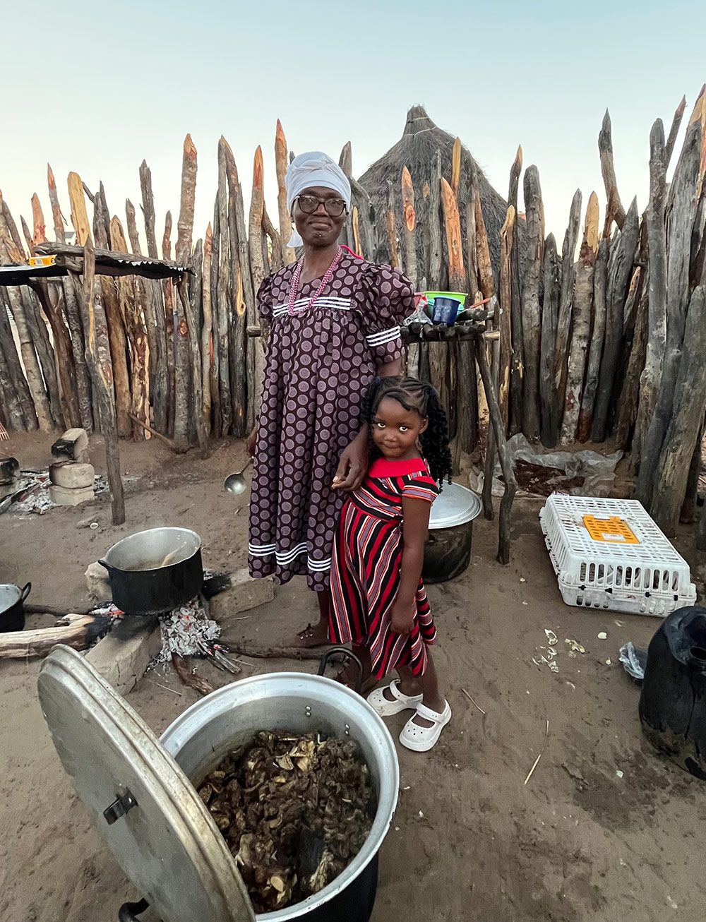 An elderly woman in a patterned dress and headscarf stands next to a young girl in a striped dress, both smiling in a rustic outdoor kitchen surrounded by wooden fencing. Cooking pots are visible in the foreground.
