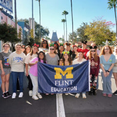 A group of people stands together in a theme park, holding a blue banner that reads "M FLINT EDUCATION." The background features palm trees and colorful buildings.
