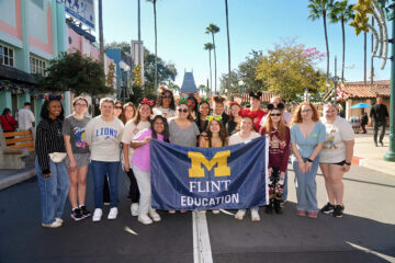 A group of people stands together in a theme park, holding a blue banner that reads "M FLINT EDUCATION." The background features palm trees and colorful buildings.